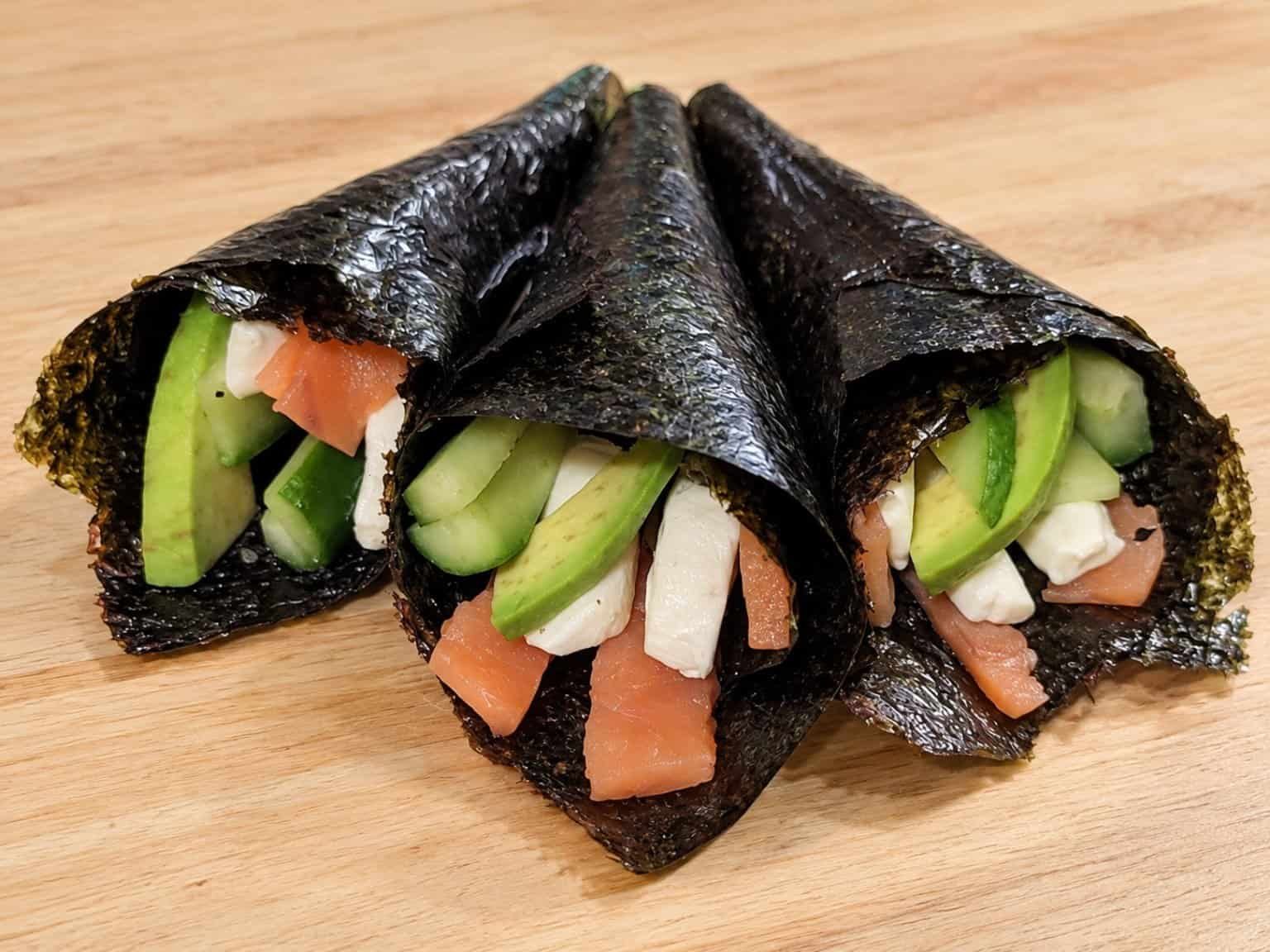 Three salmon hand rolls wrapped in nori and filled with smoked salmon, cream cheese, avocado, and cucumber.