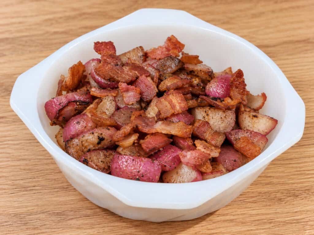 Golden-brown roasted radish pieces with red skins and crispy bacon in a white bowl on a light oak countertop.