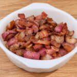 Golden-brown roasted radish pieces with red skins and crispy bacon in a white bowl on a light oak countertop.