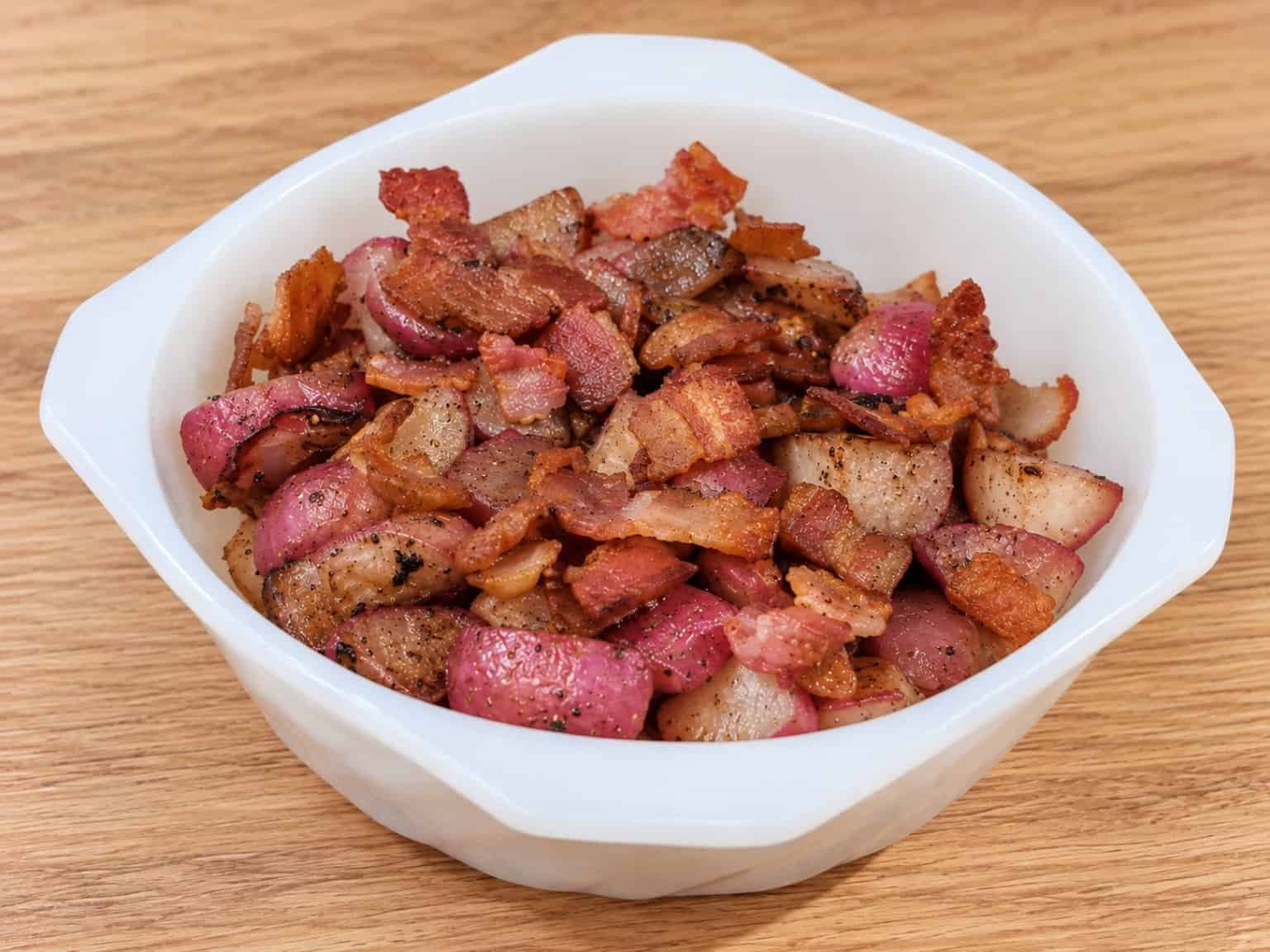 Golden-brown roasted radish pieces with red skins and crispy bacon in a white bowl on a light oak countertop.