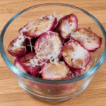 Halved garlic Parmesan roasted radishes with golden-brown surfaces, pink skins, and shredded Parmesan cheese in a clear glass bowl on a light oak countertop.