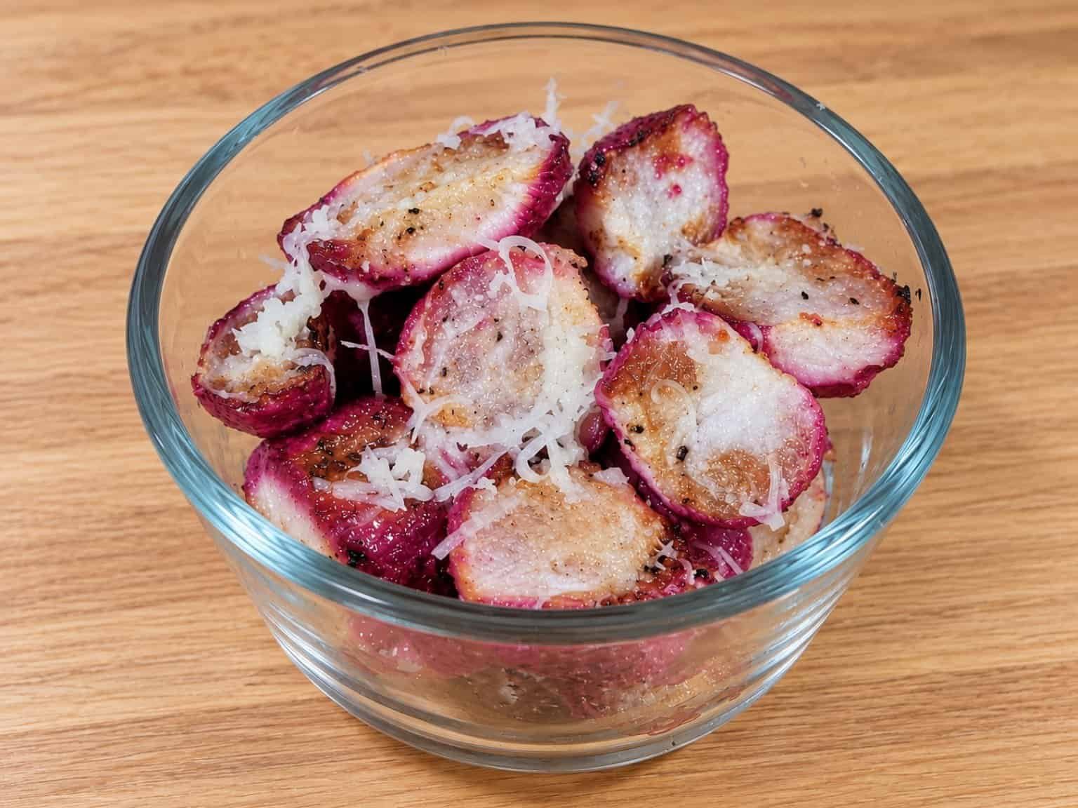 Halved garlic Parmesan roasted radishes with golden-brown surfaces, pink skins, and shredded Parmesan cheese in a clear glass bowl on a light oak countertop.
