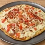 Creamy keto chicken bacon ranch pizza topped with melted cheese, diced tomatoes, chicken, and bacon crumbles on a dark gray pizza pan.