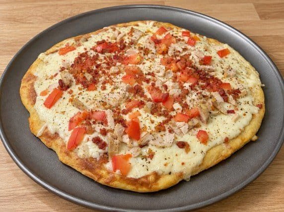 Creamy keto chicken bacon ranch pizza topped with melted cheese, diced tomatoes, chicken, and bacon crumbles on a dark gray pizza pan.